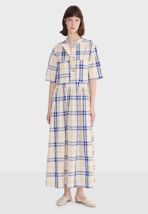 Žena stojaca v krémových šatách s modro-oranžovým károvaným vzorom, krátkymi rukávmi a béžovými nazúvacími topánkami.