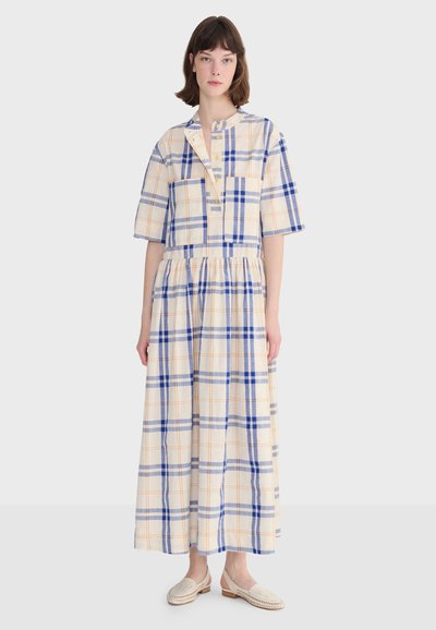 Femme debout portant une robe crème à motif écossais bleu et orange, manches courtes, et des chaussures beige à enfiler.