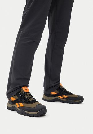 Schwarze Outdoor-Hose, getragen mit braunen und schwarzen Reebok-Trailschuhen mit orangefarbenen Akzenten und dicken, robusten Sohlen.