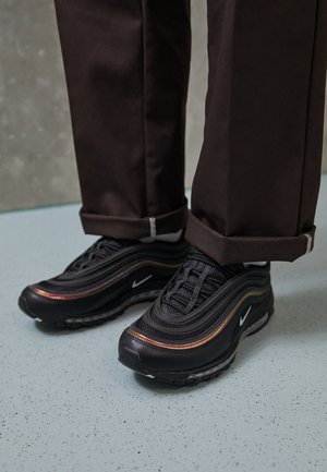 Person trägt schwarze und orangefarbene Sneakers mit weißem Swoosh-Logo und dunkelbraunen, aufgekrempelten Hosen, steht auf einem hellblauen, gesprenkelten Boden.