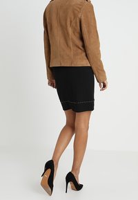 Hnědá semišová bunda v kombinaci s černou sukní po kolena, která má jemně vystouplý lem s hroty, a černými lodičkami na vysokém podpatku.