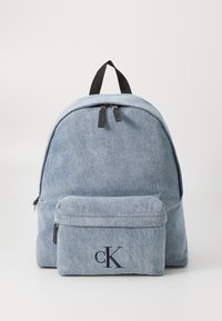 Sac à dos en denim bleu clair avec deux fermetures éclair, une poche avant et un logo "cK" en bleu marine. Texture lisse, forme arrondie, bretelles réglables.