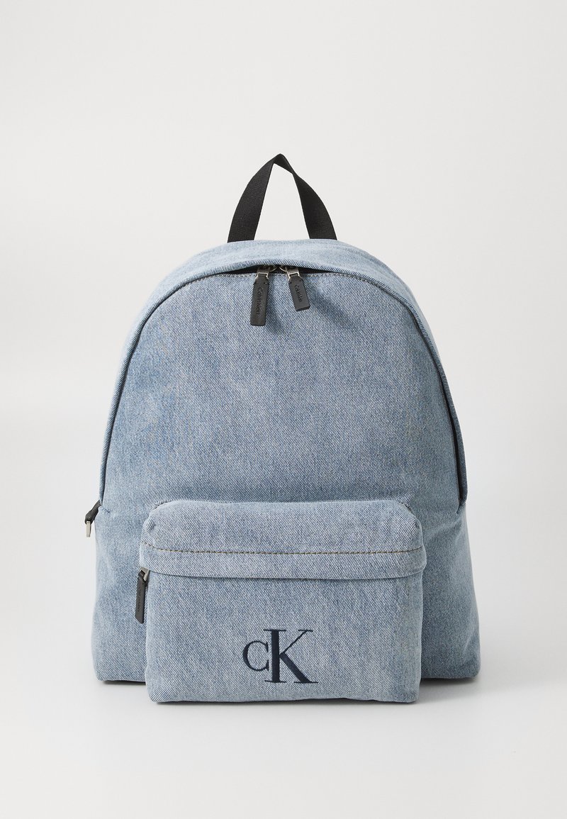 Sac à dos en denim bleu clair avec deux fermetures éclair, une poche avant et un logo "cK" en bleu marine. Texture lisse, forme arrondie, bretelles réglables.