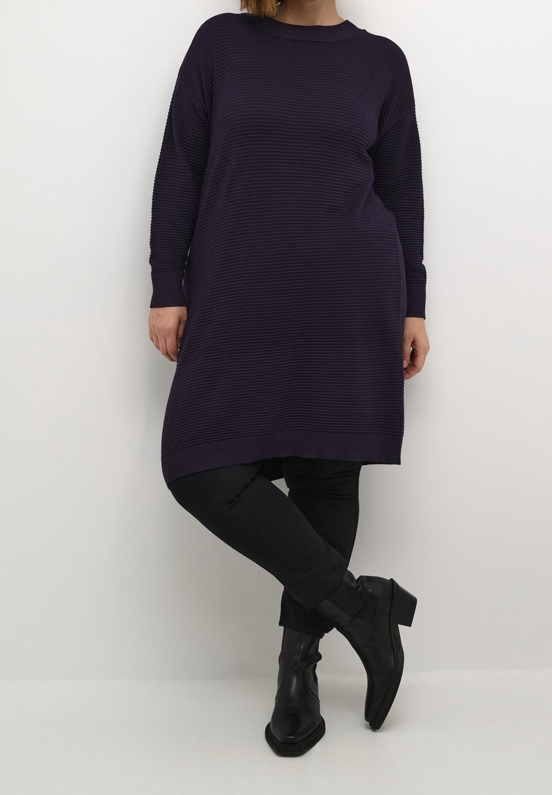 Femme portant une robe pull longue côtelée bleu marine foncé, des leggings noirs et des bottines noires, debout devant un fond clair uni.
