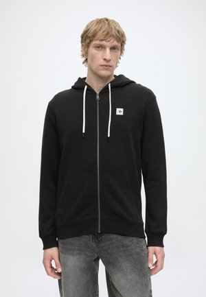 Jonge man die een zwarte hoodie met rits en witte trekkoorden draagt en een grijze spijkerbroek, staand tegen een effen witte achtergrond.