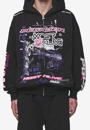 Sweat à capuche zippé noir avec texte graphique audacieux « Most Alive » en rose, blanc et violet, incluant des caractères urbains abstraits et japonais.