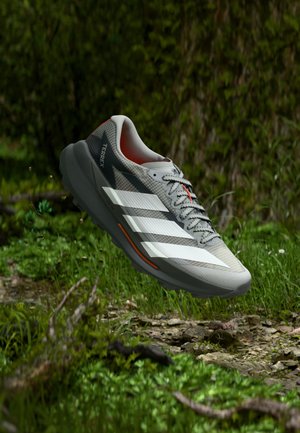 Zapatilla de trail running Adidas Terrex de color gris claro con franjas blancas y detalles en naranja, suspendida sobre suelo boscoso con hierba y rocas.