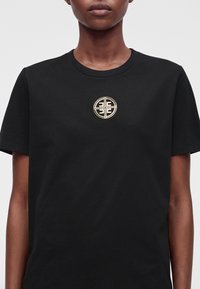 Personne portant un t-shirt noir avec un emblème rond doré et noir centré sur la poitrine montrant un motif géométrique symétrique.