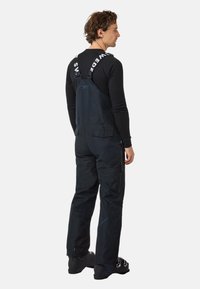 Zwarte waterdichte bretels met verstelbare banden, zijzakken met rits en verstevigde knieën, gedragen over een zwart langemouwenshirt.