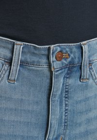 Taille d'un jean en denim avec bouton marron, passants de ceinture, coutures orange, porté avec une chemise noire rentrée.