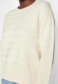 Suéter color crema con rayas horizontales texturizadas, un dobladillo acanalado y un escote redondo, que muestra un material de punto suave.