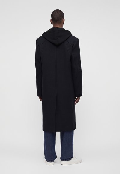 Homme debout de dos, portant un long manteau noir à capuche, un jean bleu et des baskets blanches sur un fond clair uni.