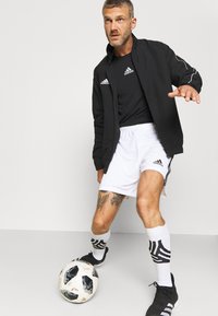 Giacca leggera nera, t-shirt Adidas nera, pantaloni corti bianchi, calze bianche fino al ginocchio con motivo nero, e scarpini da calcio neri. Palla da calcio ai piedi.