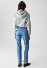 Persona con capelli corti che indossa un felpa grigia chiara, jeans blu a gamba dritta e scarpe nere, in piedi di spalle su uno sfondo neutro.