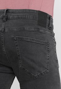 Jeans de denim negro con un corte ajustado, costuras visibles, un bolsillo trasero rectangular y una etiqueta de tela negra texturizada en la cinturilla.