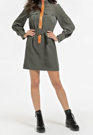 Kobieta ubrana w oliwkowo-zieloną, długorękawową mini sukienkę z pomarańczowym zamkiem błyskawicznym i paskiem, w parze z czarnymi, sznurowanymi botkami za kostkę.