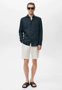 Camisa de lino de manga larga en azul oscuro, pantalones cortos blancos y sandalias negras. Hombre con gafas de sol, manos en los bolsillos, de pie contra un fondo liso.