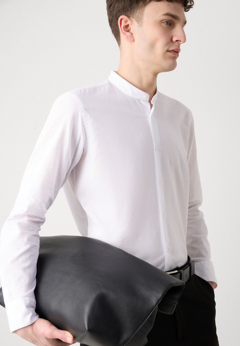 Biała, długa koszula z mandaryńskim kołnierzykiem, teksturowana tkanina, oraz czarna skórzana torba trzymana w jednej ręce na neutralnym tle.