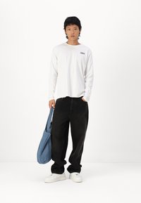 Long-sleeve white cotton shirt with a small logo, paired with loose black denim pants and white sneakers. A blue tote bag is carried.
