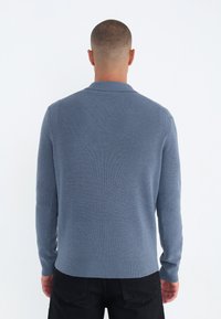Suéter de punto acanalado azul con mangas largas y cuello, que presenta un acabado texturizado y un dobladillo ligeramente entallado. Se llevan pantalones negros.