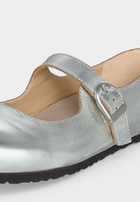 Ασημένιο παπούτσι Mary Jane με ρυθμιζόμενο λουρί με πόρπη και μαύρη σόλα, με γυαλιστερό δερμάτινο επάνω μέρος και επενδεδυμένη μπεζ εσωτερική επένδυση.