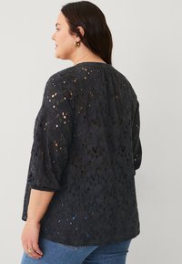 Blusa nera ricamata con ritagli floreali, maniche a tre quarti e scollo rotondo, abbinata a jeans blu. Il tessuto testurizzato aggiunge dettaglio.