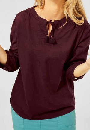 Vrouw draagt een donkerpaarse blouse met lange mouwen, een tassel strikhalssluiting en lichtblauwe broek tegen een effen achtergrond.