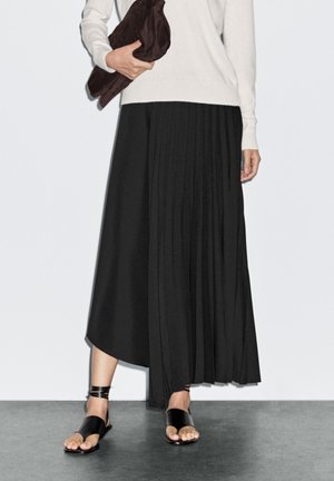 Zwarte geplooide maxi rok met een vloeiende uitstraling, gecombineerd met zwarte platte sandalen en een donkerbruine clutch, tegen een neutrale achtergrond.