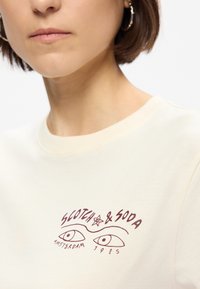 Κρεμ T-shirt με στρογγυλή λαιμόκοψη. Διαθέτει κόκκινο γραφικό σχέδιο με δύο μάτια, "SCOTCH & SODA," "ΑΜΣΤΕΡΝΤΑΜ," και "1985." Λεπτή υφή.