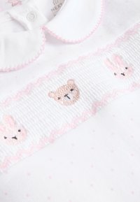 Vêtement en coton blanc à pois roses, doté d'un col festonné et de dessins brodés d'ours et de lapin sur une bande texturée.