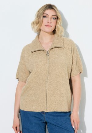 Blond vrouw met een beige kortemouwen sweater met rits en blauwe spijkerbroek, staand tegen een witte achtergrond.