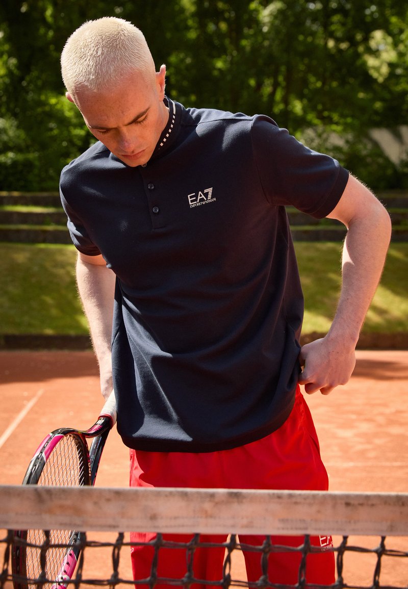 Navy polo shirt with short sleeves and a buttoned placket, featuring a logo on the chest, worn with red shorts. Holding a tennis racket.