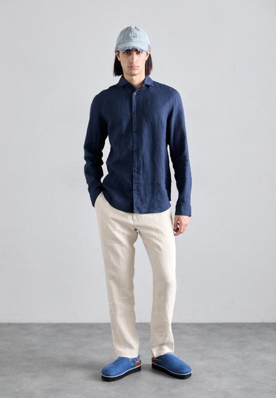 Camisa de lino azul marino con botones, pantalones de lino beige, zapatos de tela azul con suelas negras y una gorra de béisbol azul claro. Diseño simple y moderno.