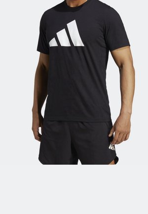 Homme portant un t-shirt noir Adidas avec un grand logo blanc et un short noir avec un petit logo blanc Adidas sur la jambe gauche.