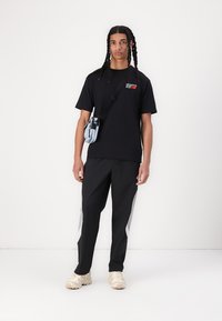 T-shirt noir en coton avec logo rouge, associé à un pantalon de survêtement noir avec des panneaux latéraux gris. Le mannequin tient un petit sac crossbody bleu clair.