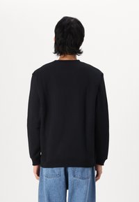 Felpa nera con collo a giro e spalle scese, con maniche lunghe e una superficie liscia. Indossata con jeans azzurri chiari dalla vestibilità ampia.