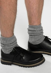 Scarpe di pelle nere con punta rotonda e lacci, abbinate a calze in maglia grigia con texture, indossate arrotolate su gambe pelose.