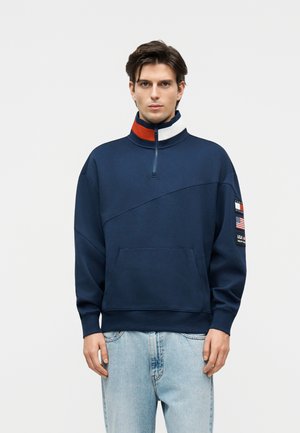 Νέος άνδρας με μπλε σκούρο φούτερ με φερμουάρ μέχρι τη μέση, με λεπτομέρειες στο γιακά σε κόκκινο, άσπρο και μπλε και μπαλώμα με σημαίες στο αριστερό μανίκι, συνδυασμένος με ανοιχτόχρωμο τζιν.