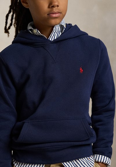 Jaunas asmuo dėvi tamsiai mėlyną su gobtuvu džemperį ant mėlynos ir baltos dryžuotos marškinių su sagomis, ant krūtinės matosi raudonas siuvinėtas logotipas.