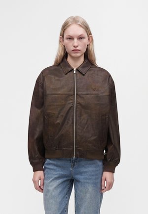 Jeune personne aux cheveux blonds longs portant une veste en cuir marron foncé avec fermeture éclair et un jean bleu clair, debout devant un fond uni.
