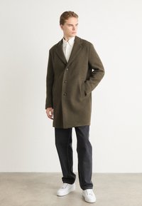 Olivgrön ullkappa med slagkrage, enkelknäppt design, tre knappar, två sidfickor, matchad med mörka jeans och vita sneakers.
