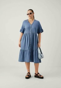 Vestido de ganga com mangas curtas e franzidas, com fecho de botões, saia em camadas, na cor azul clara. Combinado com sandálias pretas e uma pequena bolsa azul clara.