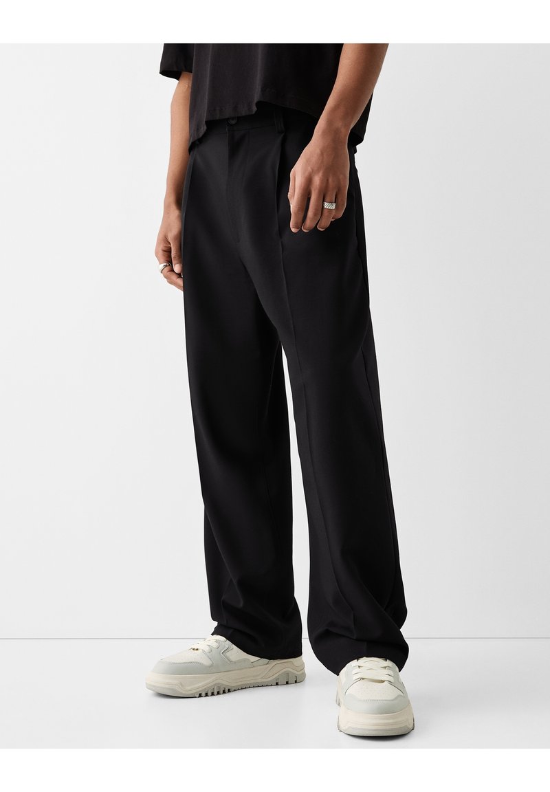 Personne portant un pantalon noir à jambes larges, un haut noir, des baskets blanches et des bagues argentées à la main gauche sur un fond clair uni.