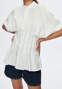 Bílá lněná halenka s krátkými nabíranými rukávy, zapínáním na knoflíky vpředu, plisovaným designem a vysokým límcem, kombinovaná s tmavými džínovými šortkami.