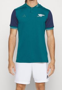 Polo bleu sarcelle avec manches bleu marine, col à deux boutons et logo brodé. Il présente des surpiqûres blanches et un tissu lisse et léger.