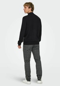 Maglione nero con collo rotondo, abbinato a pantaloni grigi a motivo check e sneakers bianche. La texture appare lavorata a maglia e liscia.