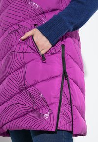 Fialová péřová vesta s lesklým vlnitým vzorem, se zipovými kapsami a černými akcenty, nošená přes modrý svetr. Viditelná texturovaná tkanina.