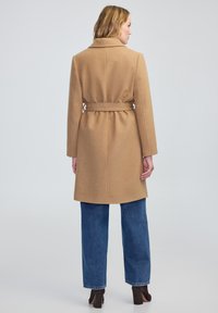 Manteau en laine couleur camel avec une ceinture, présentant un col cranté, des manches longues et une coupe mi-longue, associé à un jean taille haute.