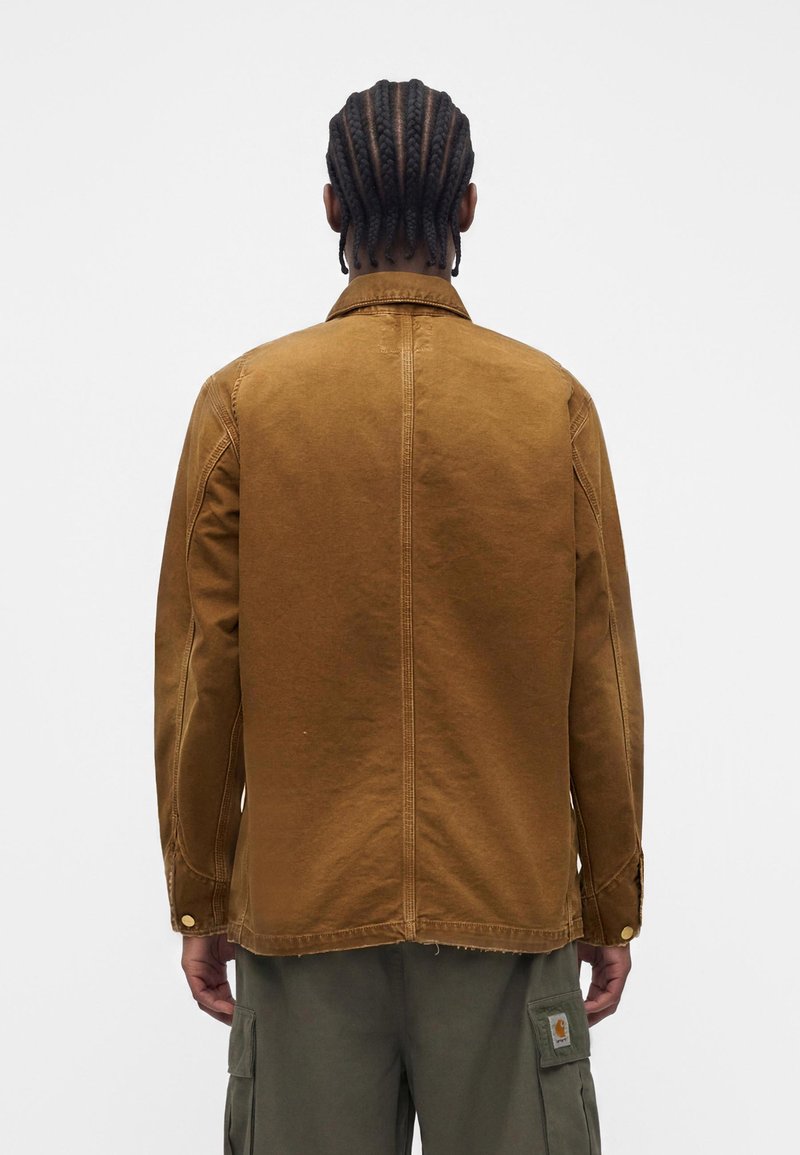 Uomo con i capelli intrecciati che indossa una giacca marrone con bottoni e maniche lunghe e pantaloni cargo color oliva, di spalle su sfondo bianco.