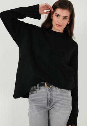 Maglione a coste nere con una vestibilità ampia e un collo alto, caratterizzato da maniche larghe e un orlo asimmetrico. Indossato con jeans grigi.